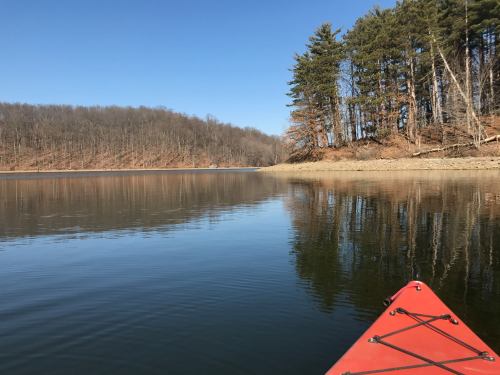 A calm lake scene with a red kayak in the foreground and trees lining the shore under a clear blue sky.