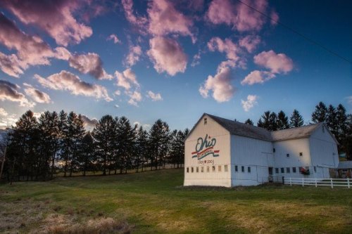 A white barn with "Ohio" painted on it, set against a colorful sky and surrounded by trees and grassy fields.