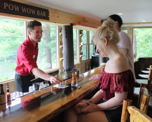A bartender in a red shirt serves drinks to a smiling woman at a rustic bar with large windows overlooking nature.