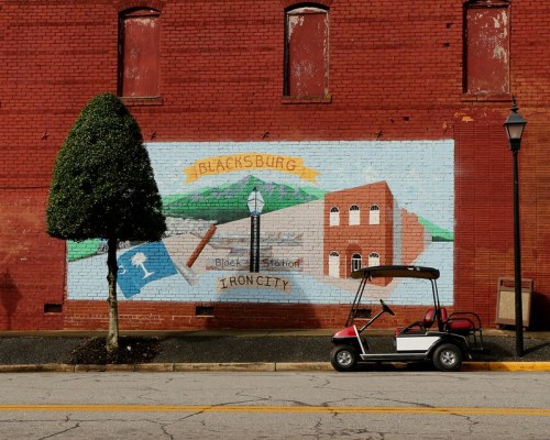 A mural on a red brick wall features a landscape, tools, and text, with a golf cart parked nearby on the street.