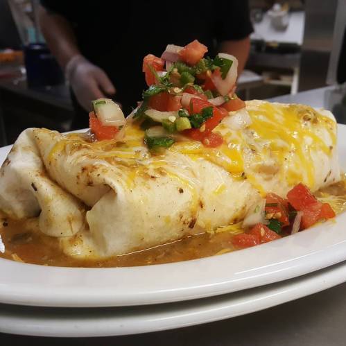 A large burrito topped with melted cheese and fresh salsa, served on a white plate in a restaurant kitchen.
