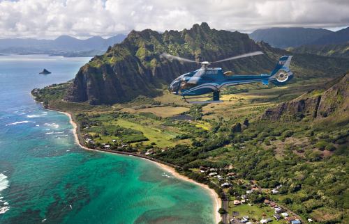 A helicopter flies over lush green mountains and a turquoise coastline, showcasing a stunning aerial view of the landscape.