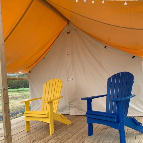 Two adirondack chairs, one yellow and one blue, sit under a large orange tent on a wooden deck.