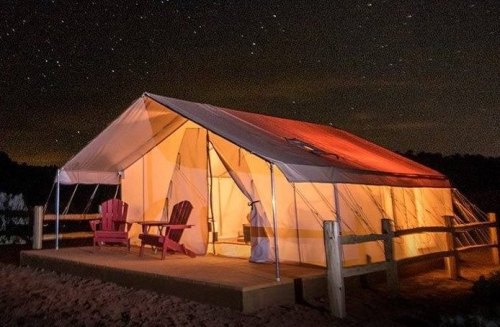 A cozy tent illuminated at night, with two chairs on a wooden deck under a starry sky.