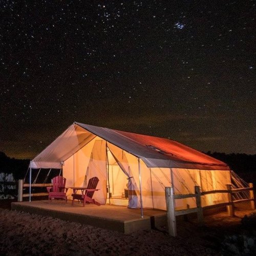 A spacious tent with glowing interior under a starry night sky, featuring two red chairs on a wooden deck.