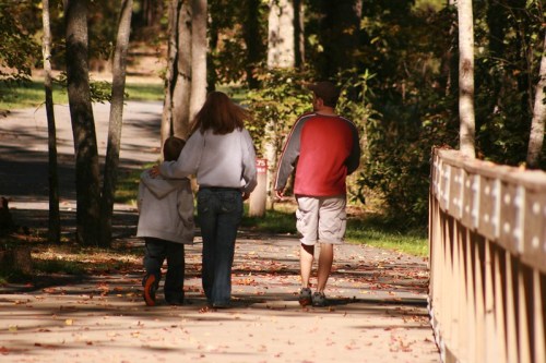 A family walks together on a tree-lined path, enjoying a sunny day in a park.
