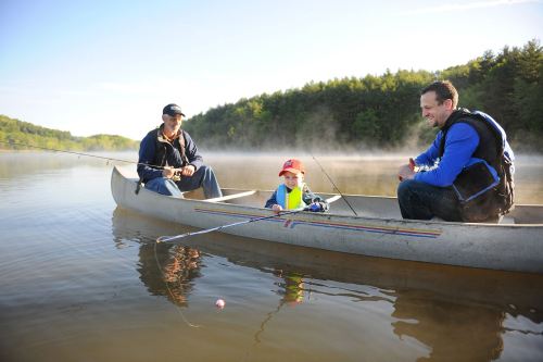 A man and a boy fish from a canoe, while another man sits nearby, surrounded by a misty lake and trees.