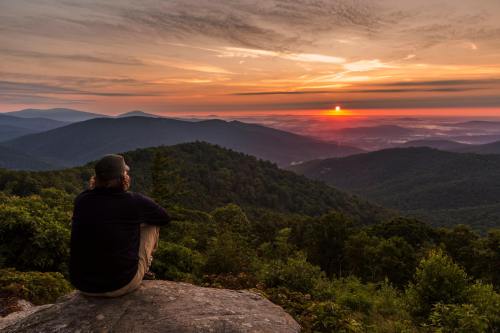 A person sits on a rock, gazing at a vibrant sunset over rolling mountains and valleys.