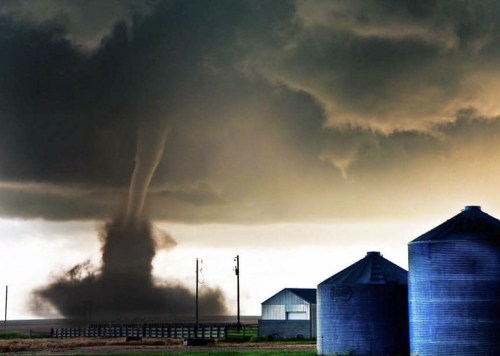A tornado forms in a stormy sky near silos and a barn, highlighting a dramatic weather scene.