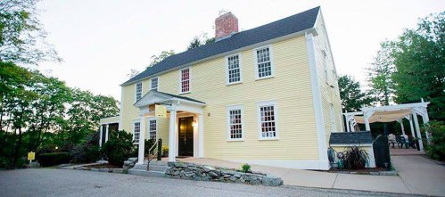 A charming yellow two-story house with white trim, surrounded by greenery and a stone pathway.