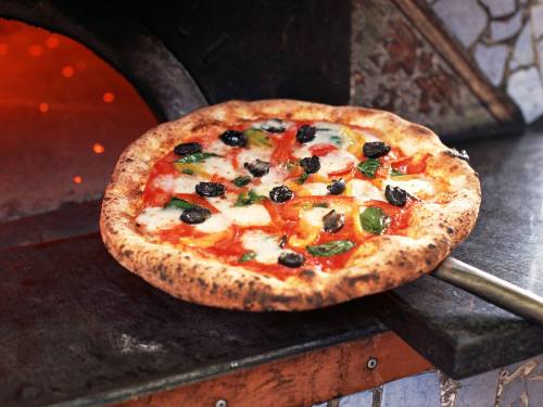 A freshly baked pizza with mozzarella, tomatoes, olives, and basil, resting on a pizza peel near a wood-fired oven.