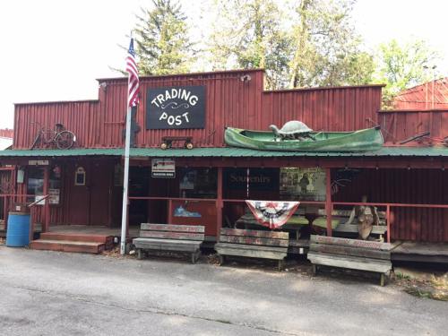 A rustic trading post with a red exterior, featuring a canoe on the roof and benches out front, surrounded by trees.