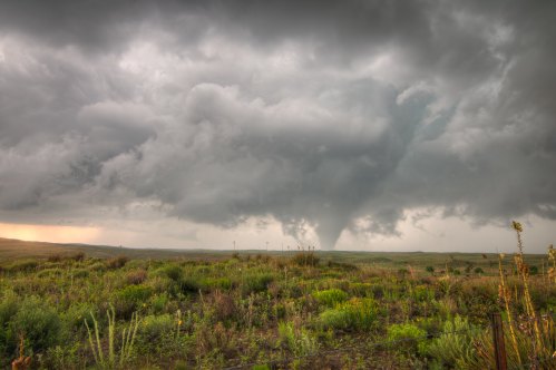 A dramatic sky with dark storm clouds and a visible tornado forming over a green landscape.