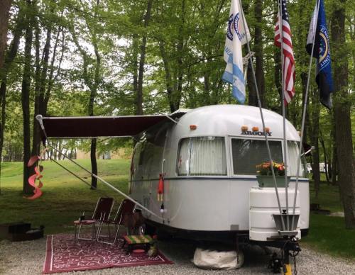 A vintage silver trailer parked in a wooded area, adorned with flags and a flower box, with chairs and a rug outside.