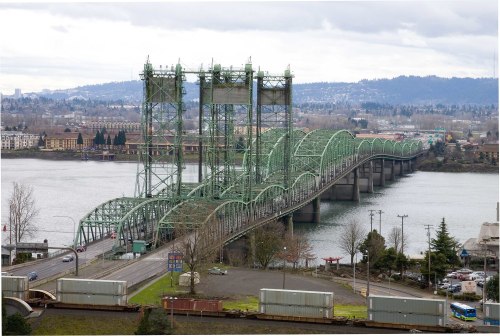 A green steel bridge spans a river, with vehicles on the road and a cloudy sky in the background.