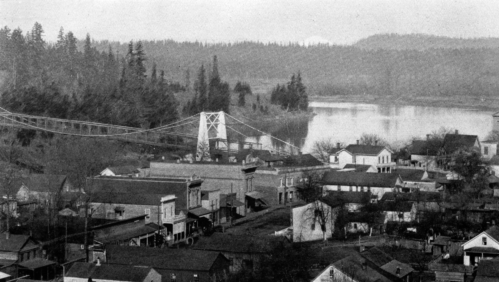 A historic black-and-white view of a small town with a suspension bridge over a river, surrounded by trees.