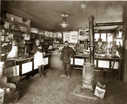 A vintage store interior with three men, shelves of goods, and a stove, showcasing early 20th-century retail.