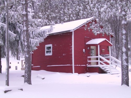 A small red cabin surrounded by snow-covered trees in a winter landscape. Snow blankets the ground and roof.