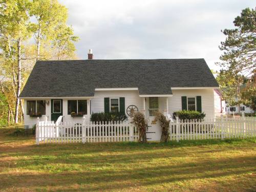 A charming white cottage with a gray roof, surrounded by a white picket fence and green lawn, under a cloudy sky.