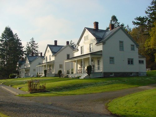 A row of white, two-story houses with porches, surrounded by green grass and trees under a clear sky.