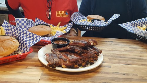 A plate of ribs with a side of beans, surrounded by burgers and fries in red baskets on a wooden table.
