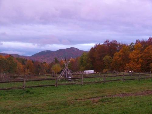 A serene landscape featuring a wooden fence, a teepee, and colorful autumn trees with mountains in the background.