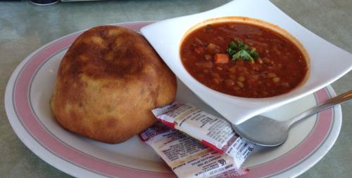 A bowl of hearty soup with vegetables, served alongside a golden-brown bread roll and condiment packets.