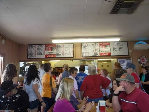 A busy diner with a crowd of people waiting in line to order at the counter, menu boards visible above.