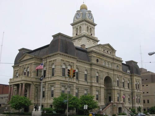 Historic stone courthouse with a clock tower, surrounded by trees and flags, under a cloudy sky.
