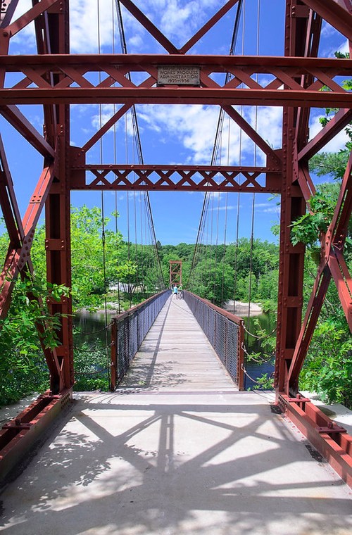 A red suspension bridge spans a river, surrounded by lush greenery and a bright blue sky with scattered clouds.