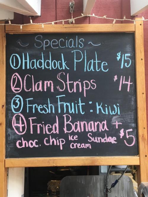 Chalkboard menu listing specials: Haddock Plate $15, Clam Strips $14, Fresh Fruit: Kiwi, Fried Banana with ice cream $5.