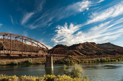 A rusted metal bridge spans a river, with rugged hills and a blue sky filled with wispy clouds in the background.