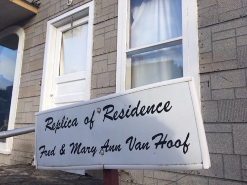 Sign reading "Replica of Residence Fred and Mary Ann Van Hoof" in front of a building with a white door and window.