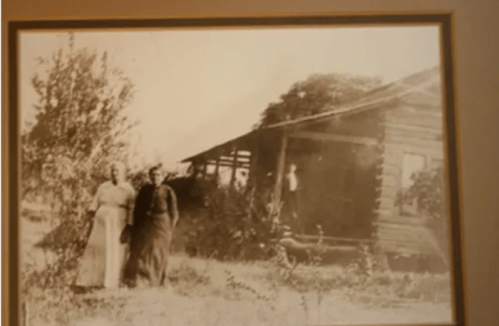 Two women stand in front of a rustic wooden house, with a figure partially visible in the background among the trees.