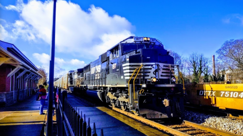 A black and white freight train passes by a station under a blue sky with scattered clouds.