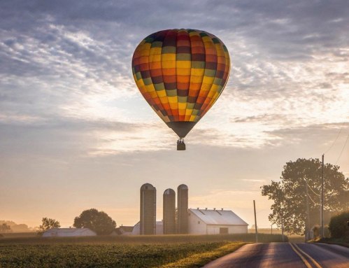 A colorful hot air balloon floats above a farm at sunrise, with silos and a winding road in the foreground.