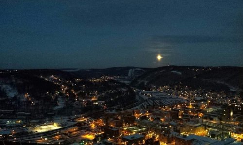 A nighttime cityscape with twinkling lights and a bright moon over snow-covered hills.