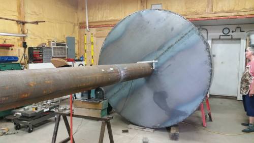 A large metal cylinder connected to a circular metal disc in a workshop, with a person observing nearby.