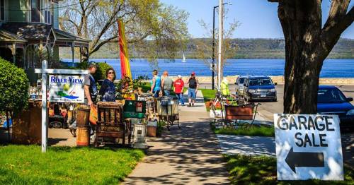 A sunny day at a garage sale by the river, with people browsing items and a sign pointing to the sale.