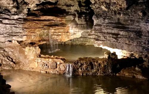 A dimly lit cave with rock formations and a small waterfall flowing into a calm pool of water.