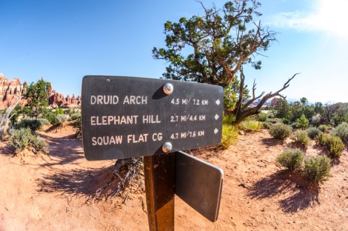 Trail sign indicating distances to Druid Arch, Elephant Hill, and Squaw Flat Campground in a desert landscape.