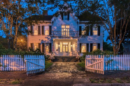 A charming two-story house with white shutters, illuminated windows, and a picket fence, set in a serene evening scene.