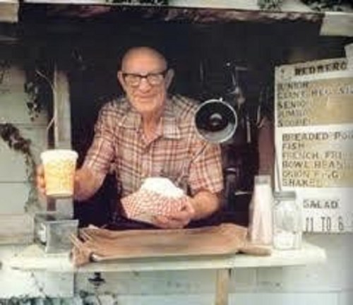 An elderly man in a plaid shirt stands at a food stall, holding a drink and a food container, with a menu in the background.