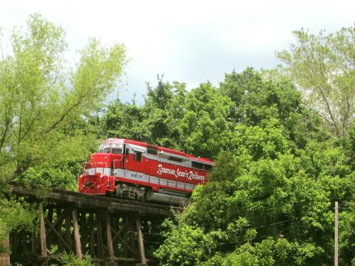 A red train on a wooden bridge surrounded by lush green trees under a cloudy sky.