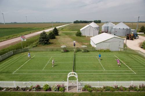 Aerial view of a tennis match on a grass court near farm buildings and wind turbines in a rural landscape.