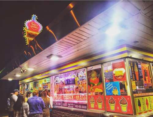 A brightly lit ice cream stand with colorful signage and a line of customers waiting outside at night.