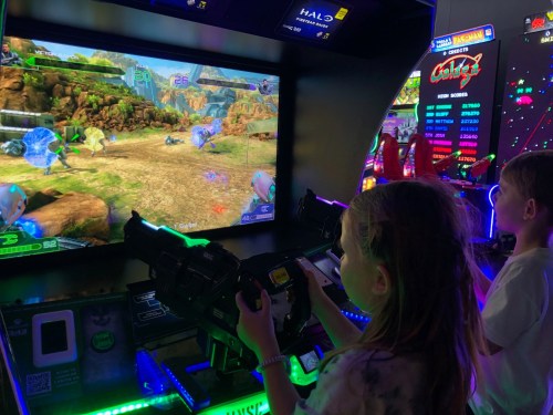 Two children play a shooting arcade game, aiming at the screen with large toy guns in a colorful gaming environment.