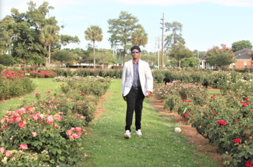 A person in a white blazer walks through a rose garden, surrounded by blooming flowers and greenery.