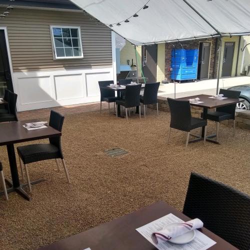 Outdoor dining area with tables set under a tent, gravel flooring, and a building in the background.