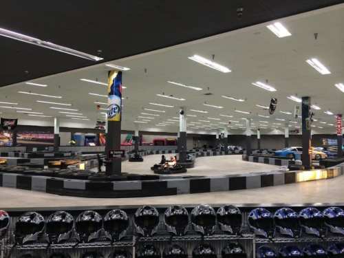 Indoor go-kart track with karts on the course and helmets displayed in the foreground. Brightly lit and spacious.